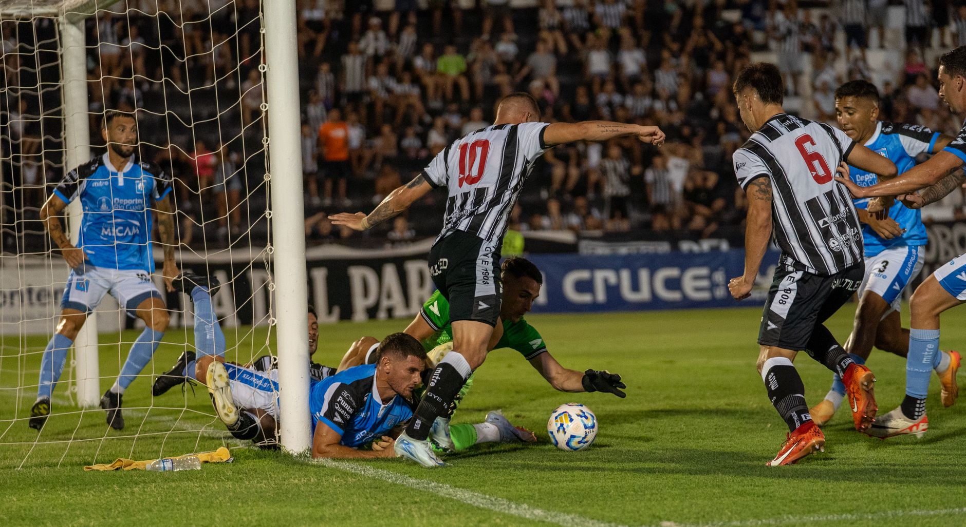 Dramatic goal-line action during a night soccer match, players in black and blue jerseys.