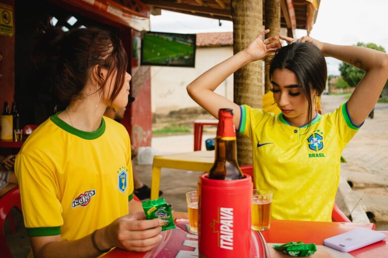 Two women in Brazil football jerseys enjoying drinks at an outdoor bar during a match.