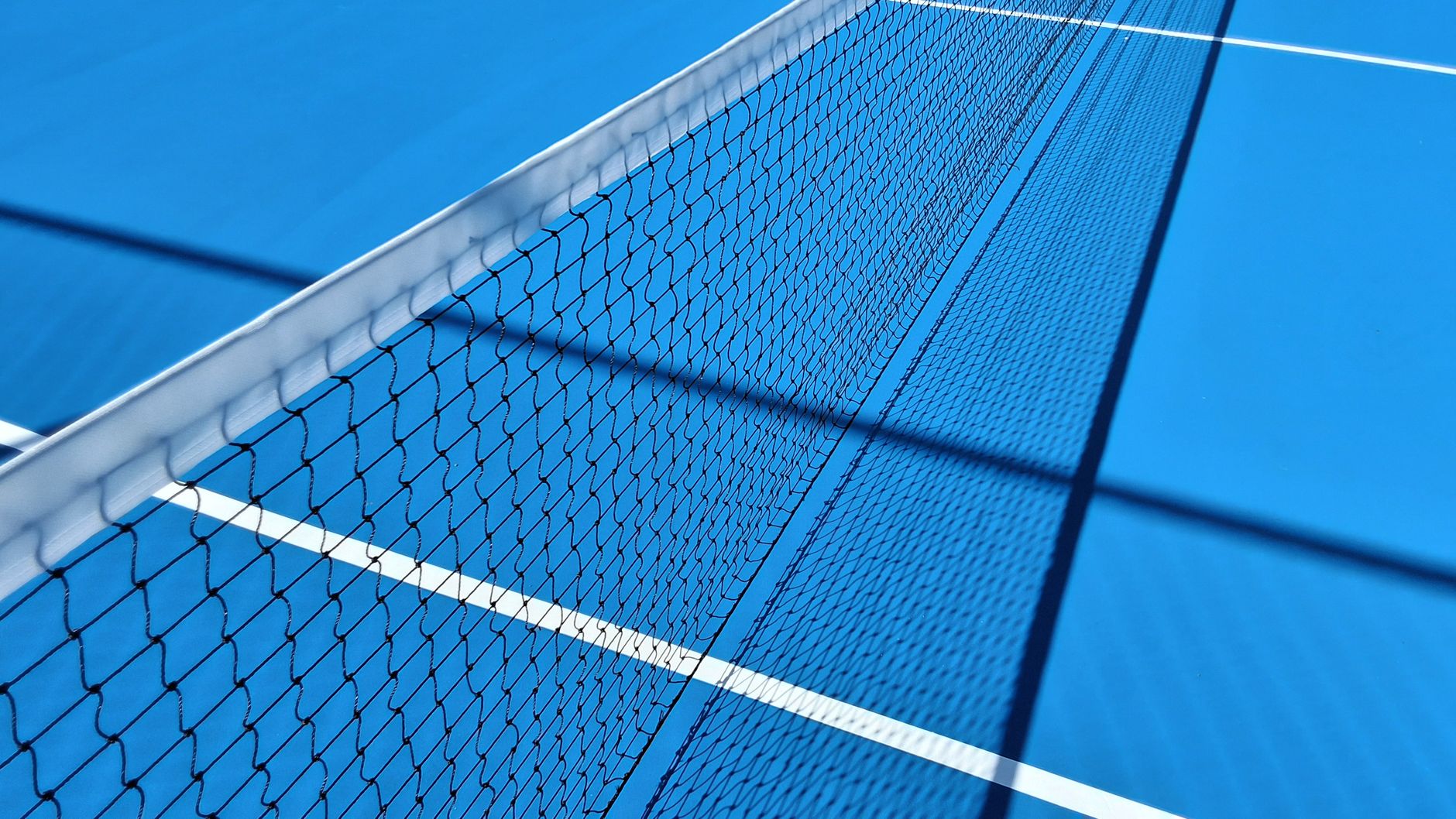 Perspective view of a blue tennis court highlighting the net and shadows under sunlight.