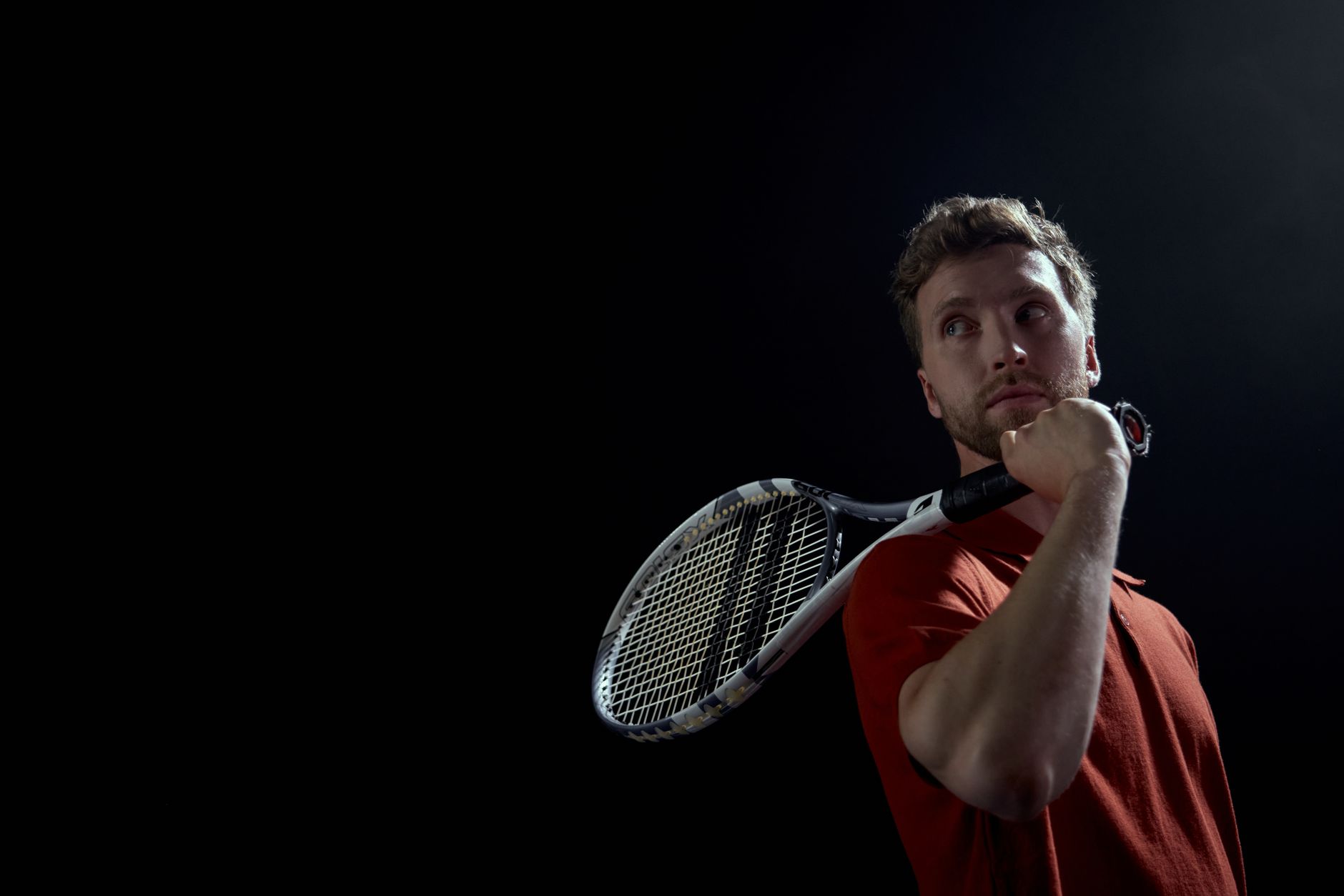 A thoughtful male tennis player holding a racket, set against a dark background.