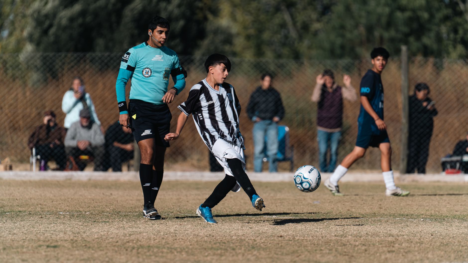A youth soccer player strikes the ball during a match with a referee nearby, expressing focus and teamwork.
