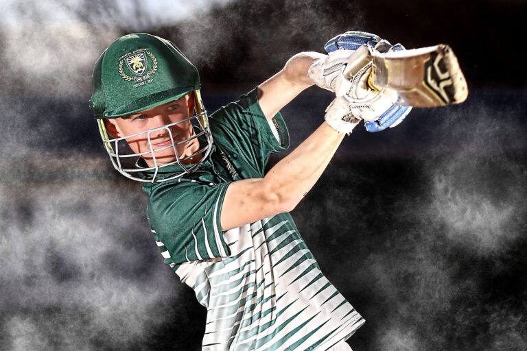 Cricket player in mid-swing with bat, wearing green uniform and helmet.