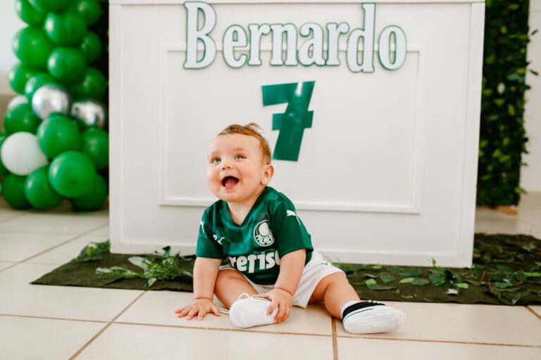 Adorable baby boy sitting indoors in a green soccer jersey, smiling happily.