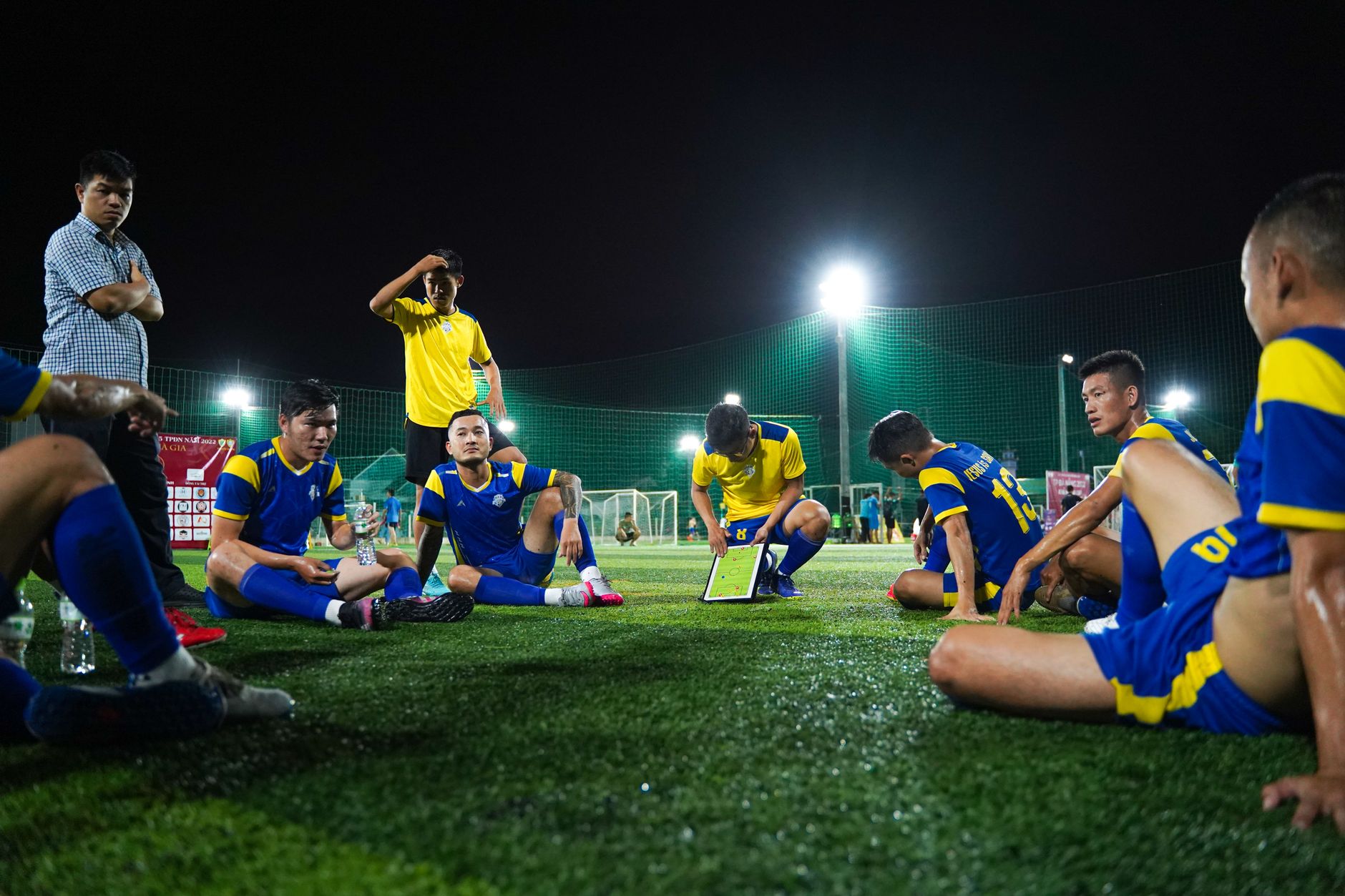 Soccer players in blue and yellow uniforms gather around coach for strategy discussion on field at night.