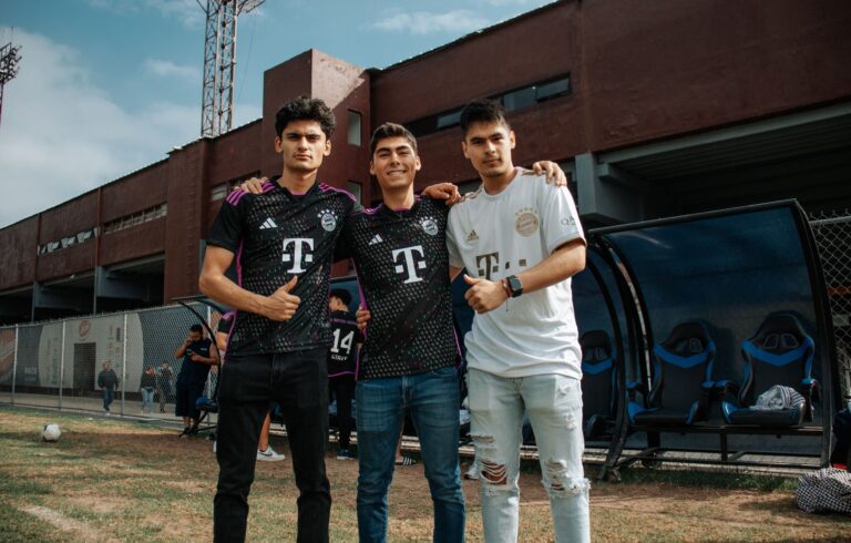 Three young men in Bayern Munich jerseys smiling at a soccer stadium, showcasing sports fandom.