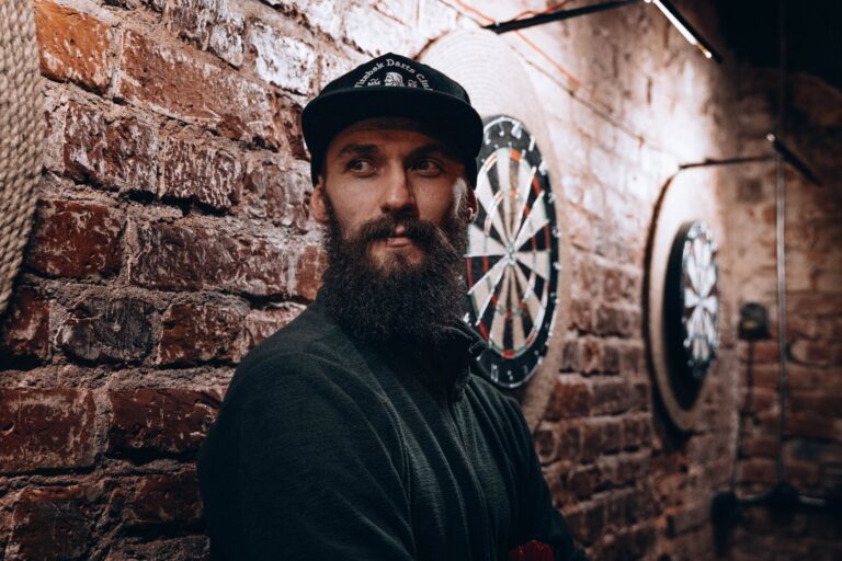 A bearded man in casual wear intently plays darts in a brick-walled setting, exuding concentration.