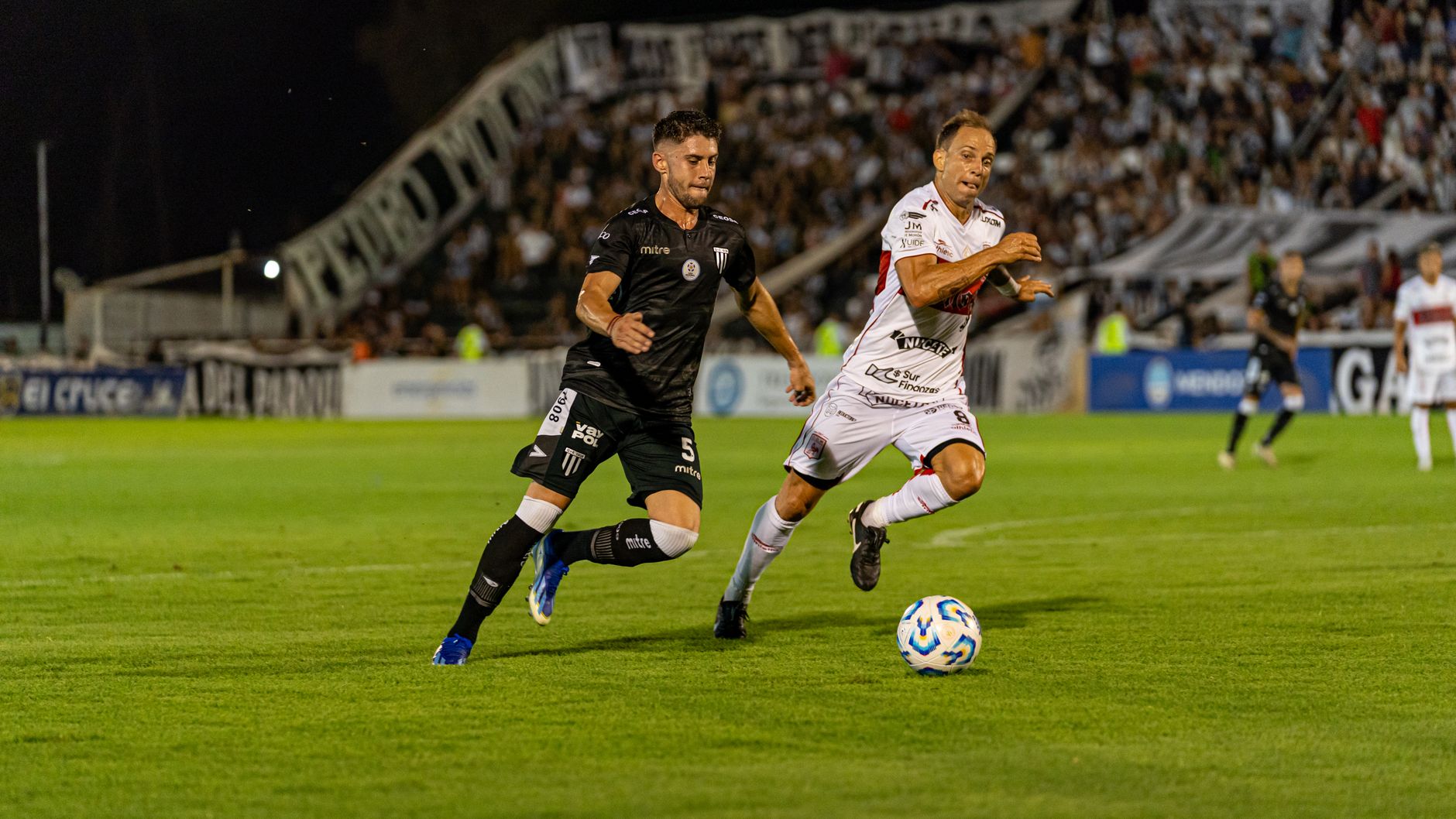 Two soccer players competing vigorously during a night match in a stadium, full crowd in background.