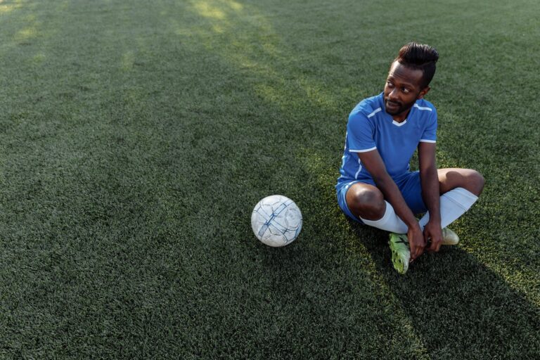 A soccer player in a blue jersey sits on the grass field during the day.