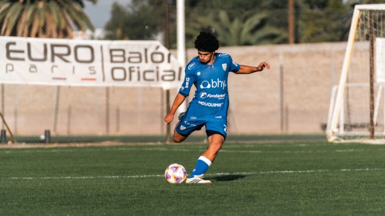 Young soccer player in blue uniform kicking a ball during a match on a sunny day.