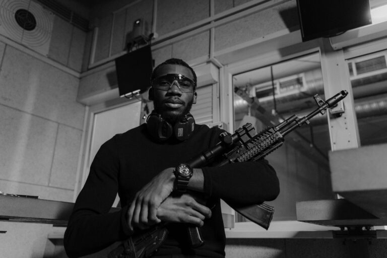 Black and white image of a man with a rifle at a shooting range, wearing safety equipment.
