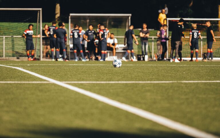 Soccer field scene with a ball in focus and players in the background during daytime.