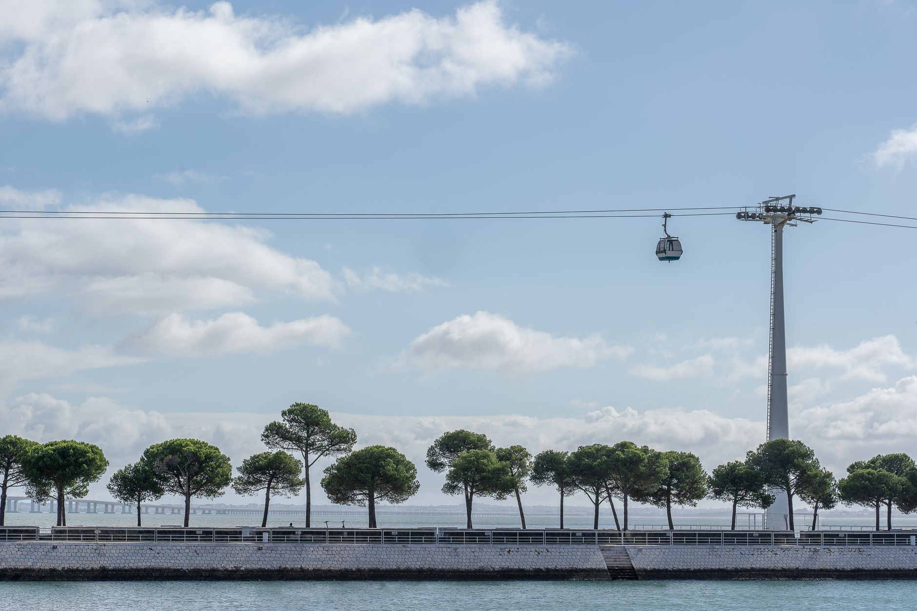 Cable car travels over tranquil seaside path lined with trees, under a clear summer sky.