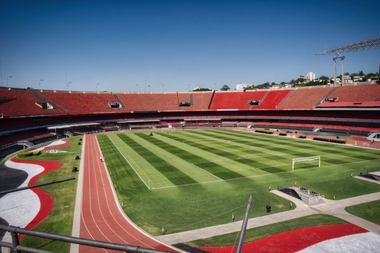 Wide shot of an empty stadium with green field and red stands under a clear blue sky.