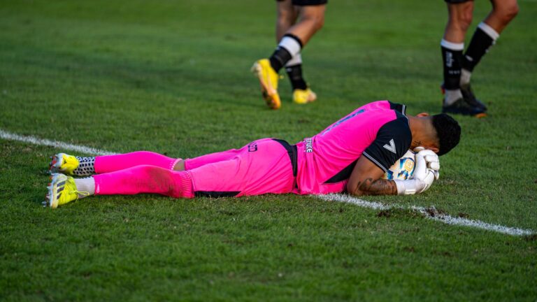 Soccer goalie in bright pink kit lying on grass field during a match.