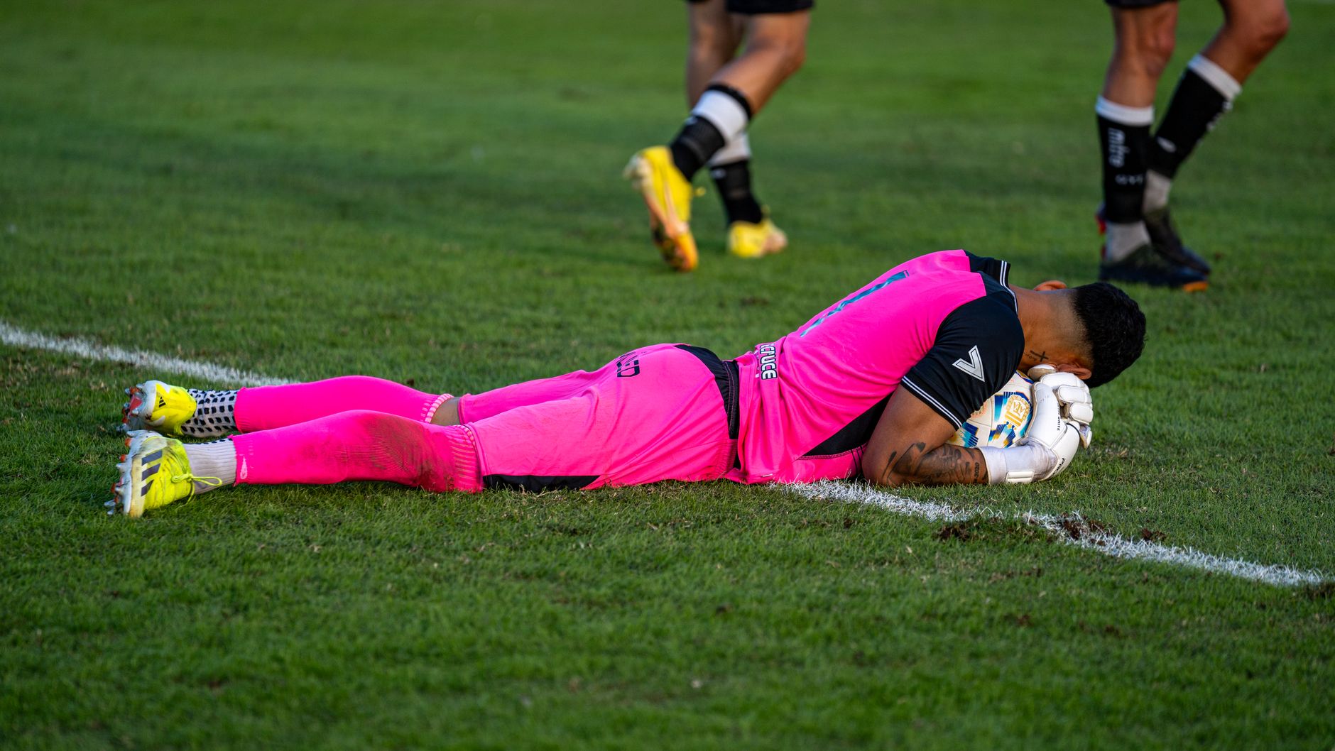 Soccer goalie in bright pink kit lying on grass field during a match.