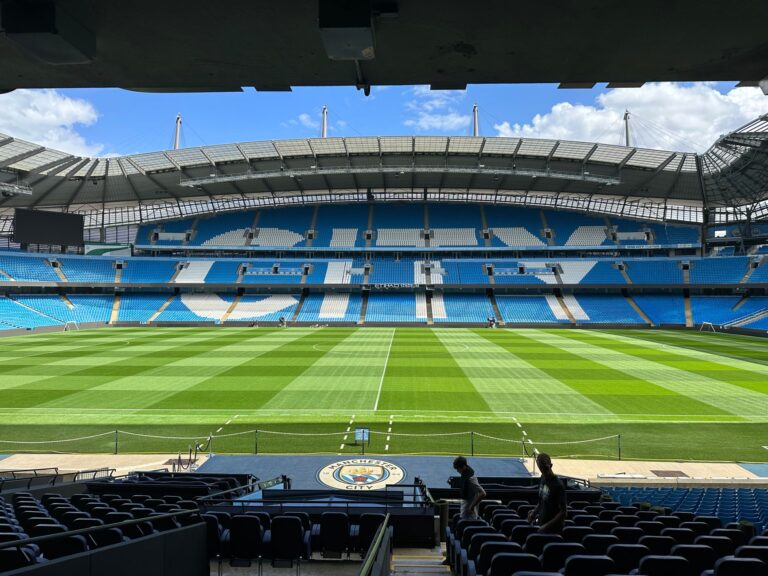 Panoramic view of Etihad Stadium, showcasing the empty football pitch and stands under a clear blue sky.