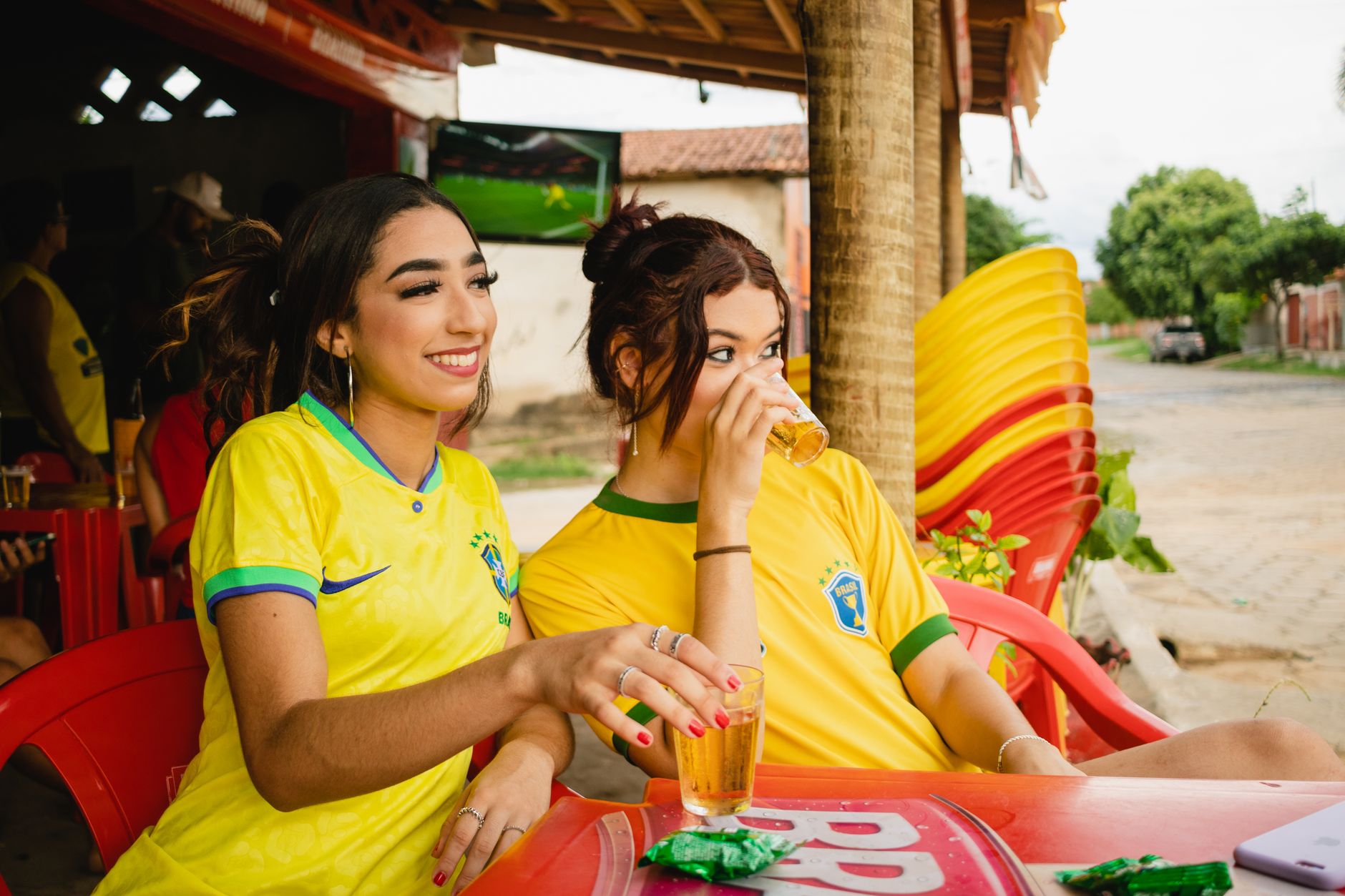 Two young women in Brazil jerseys enjoying drinks at an outdoor café, capturing summer leisure.