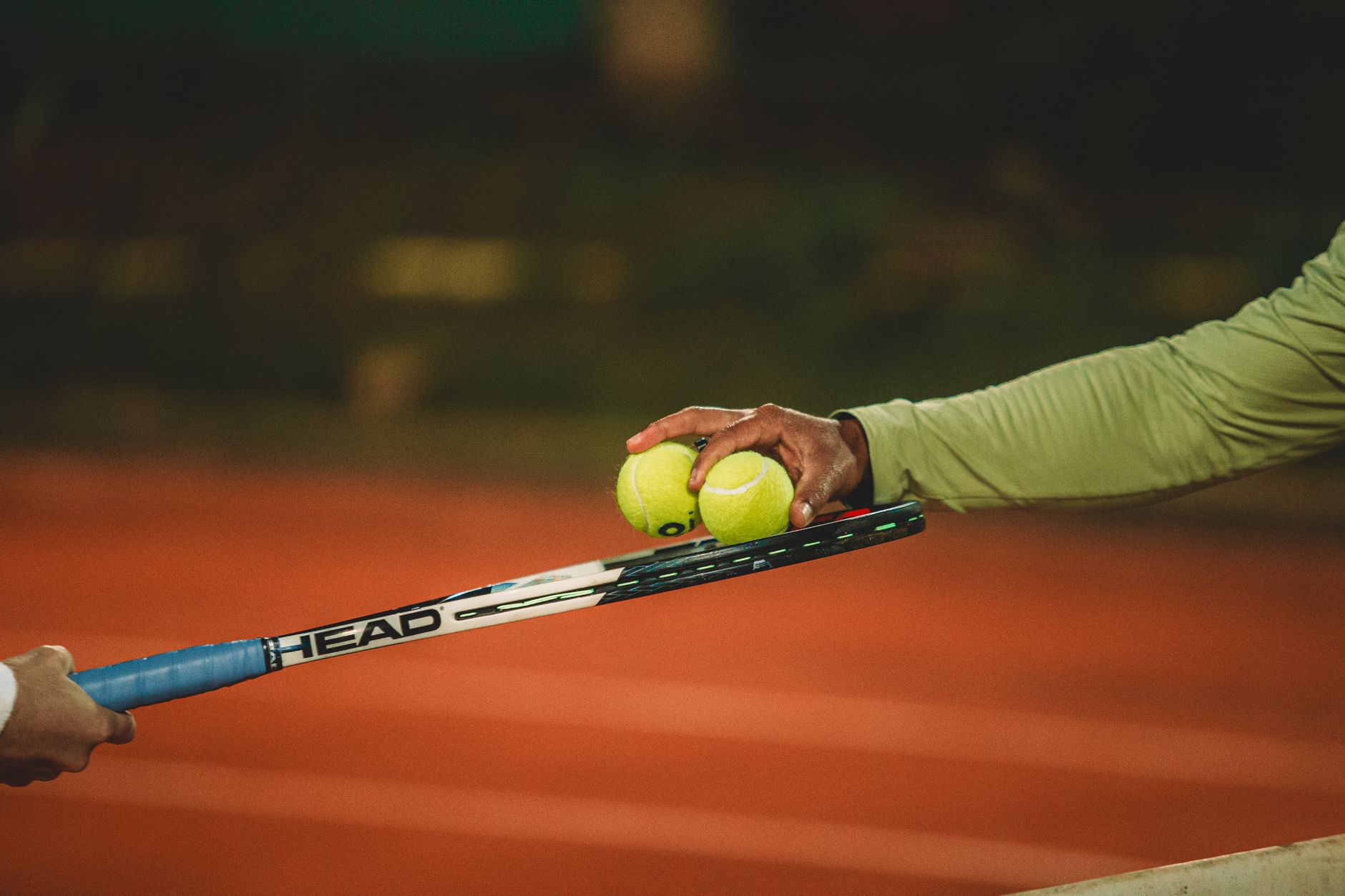 Two people exchanging tennis balls on a clay court during nighttime, focusing on the sport's dynamics.