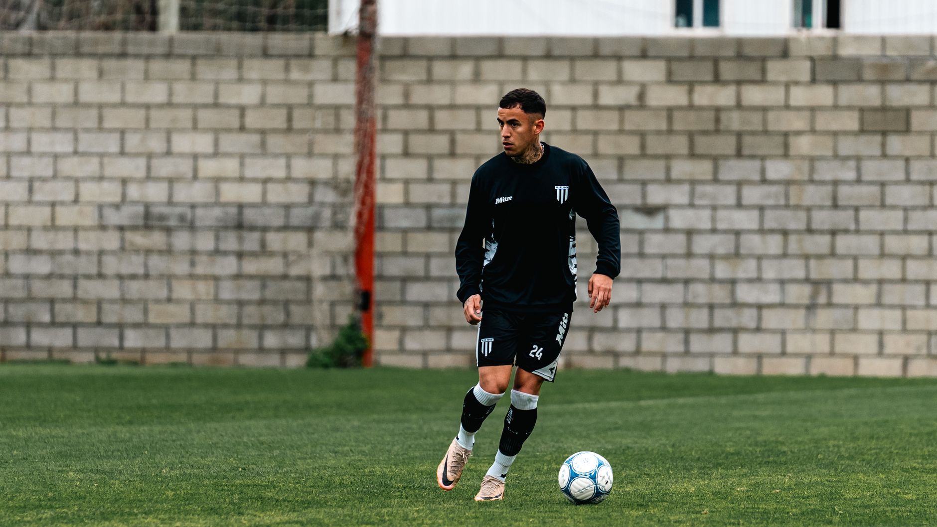 Soccer player in black kit practicing dribbling on grass field by a brick wall.
