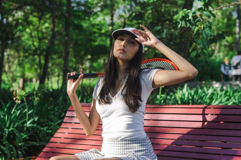 Stylish woman in casual attire posing with tennis racket on outdoor bench.