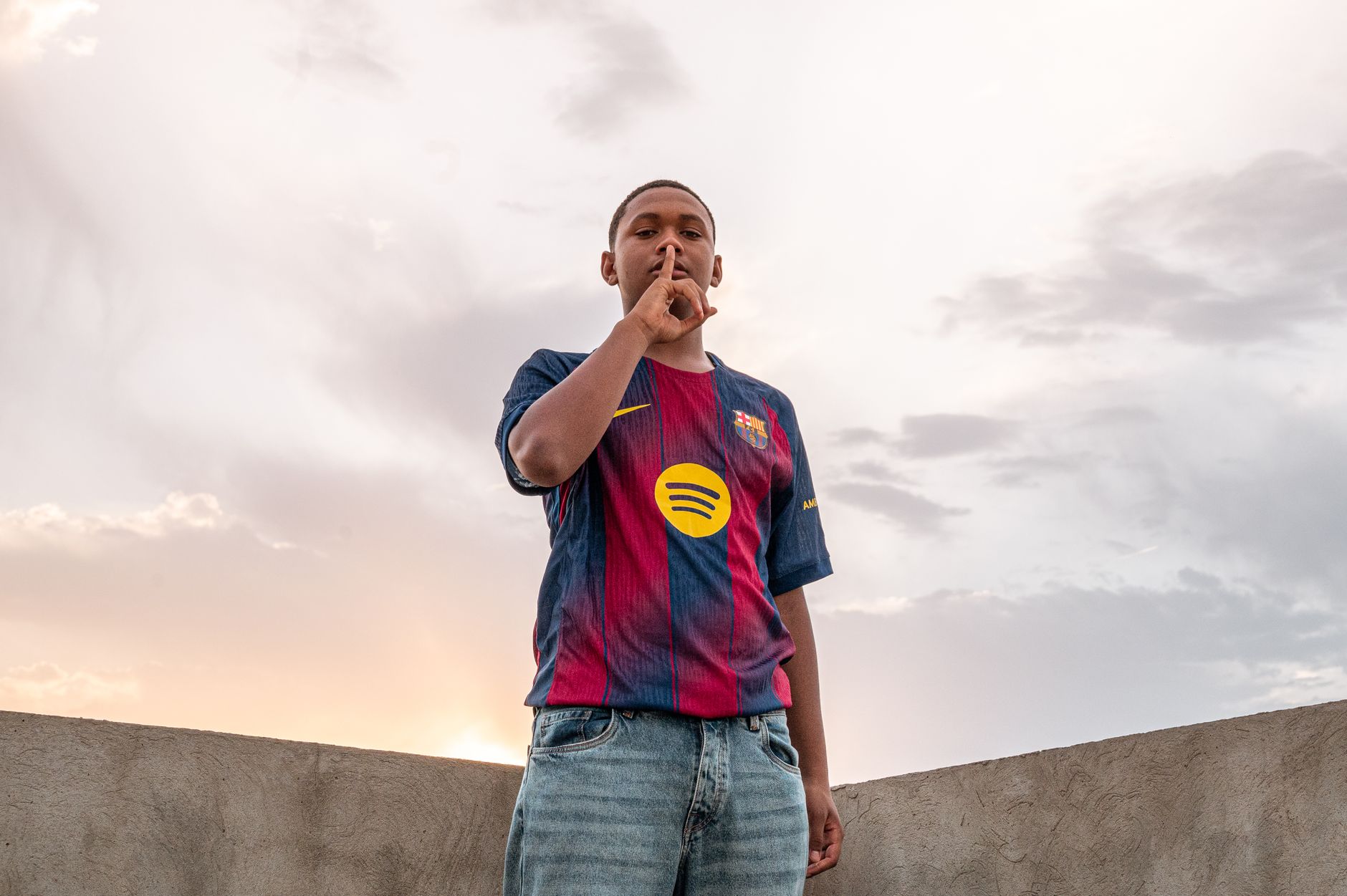 A young man in a soccer jersey signaling silence against a cloudy sky.