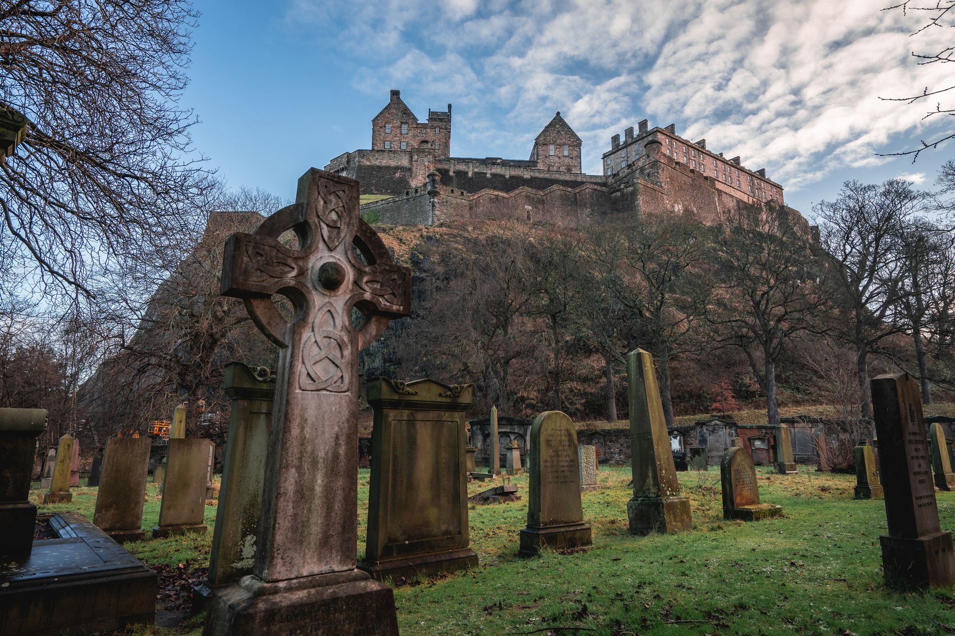 Celtic cross gravestones with Edinburgh Castle in the background, under a cloudy sky.