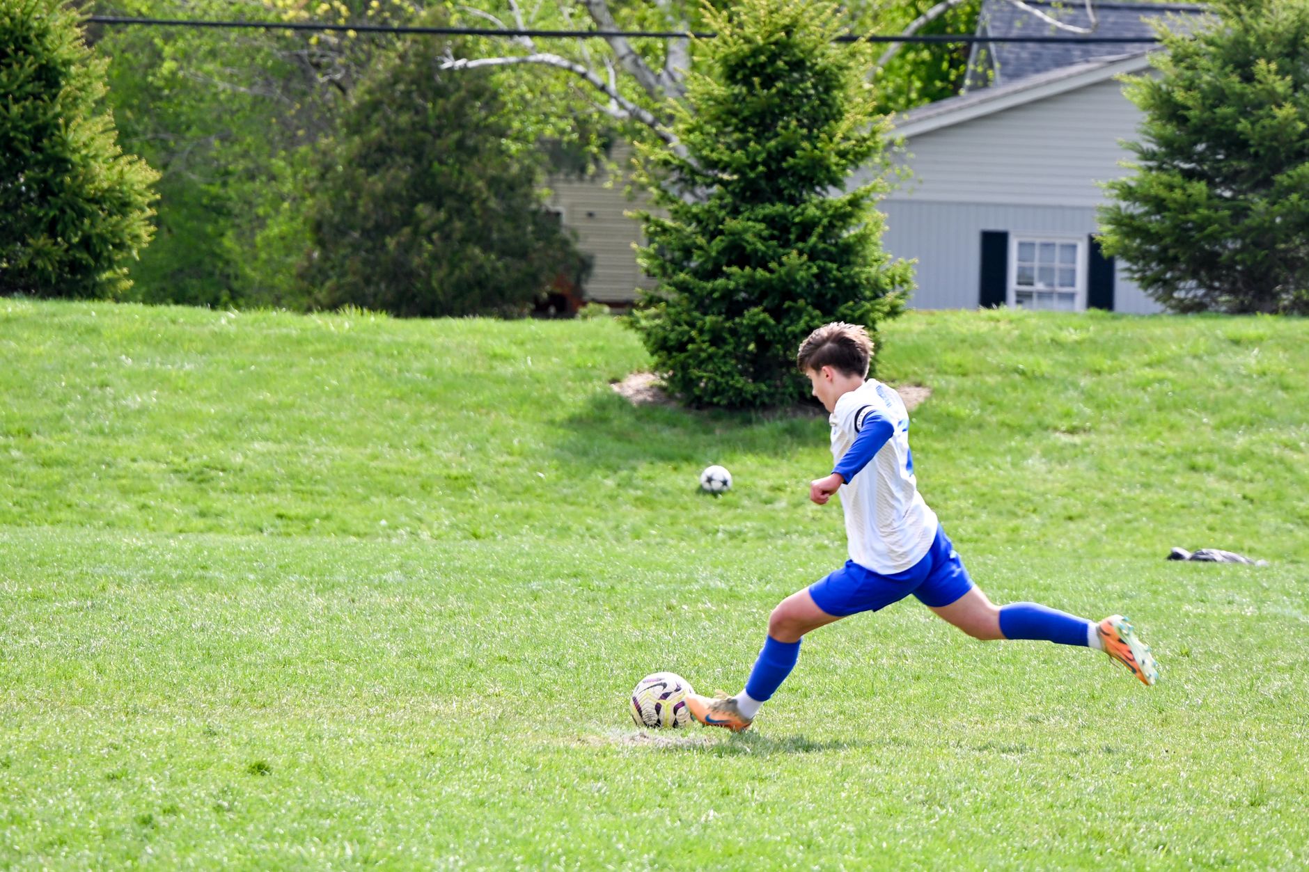 Young male soccer player in blue and white uniform kicks a ball on a grassy field outdoors.