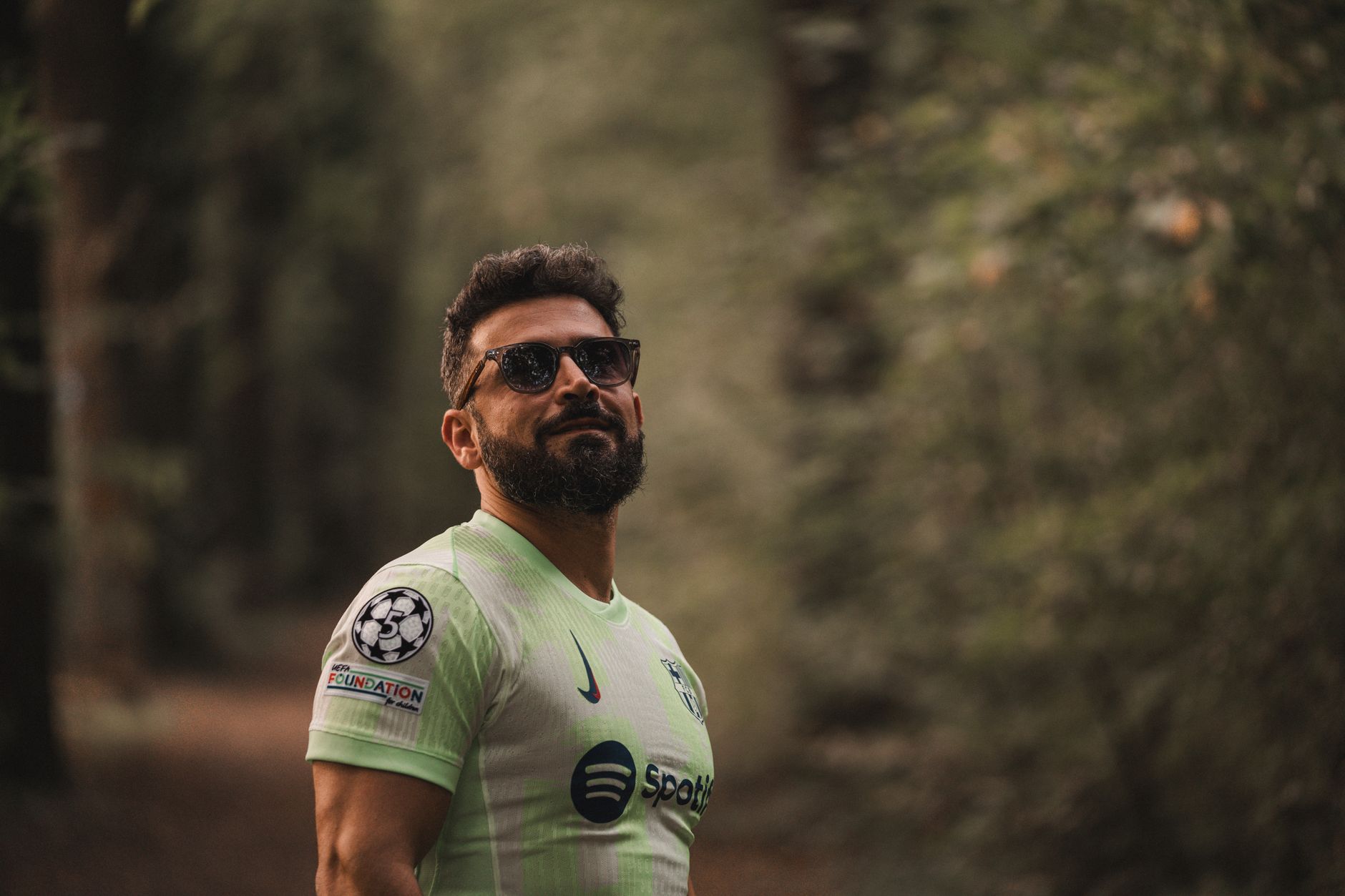 A bearded man in a soccer jersey stands confidently in a serene forest setting.