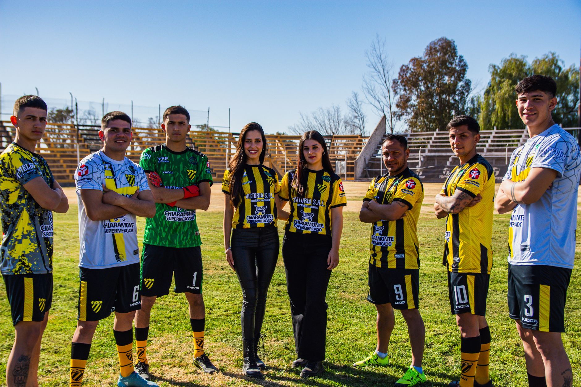 Seven soccer players and two officials in team uniforms on a sunny day.