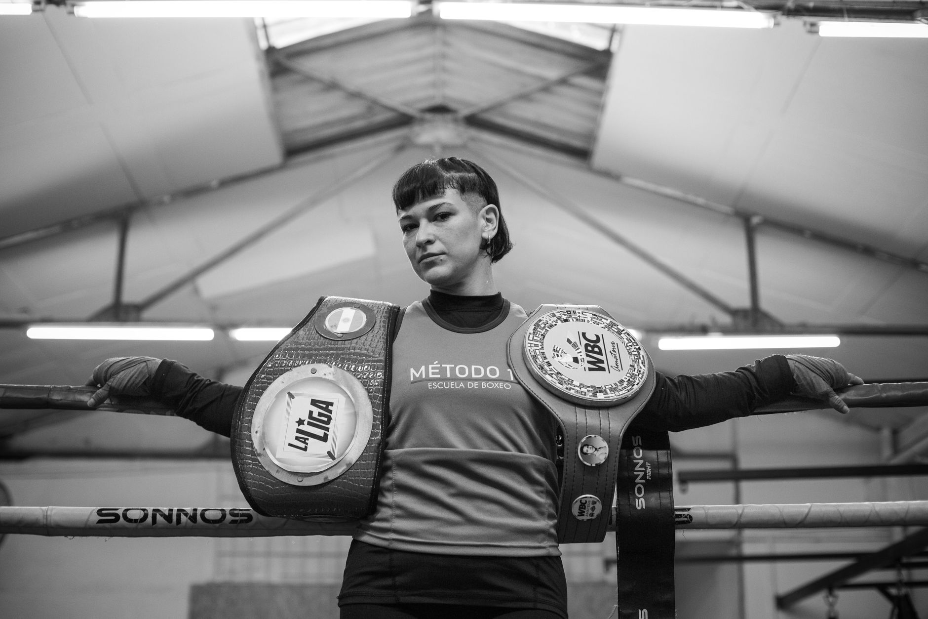A female boxer confidently poses in a gym with her championship belts, showcasing strength and achievement.