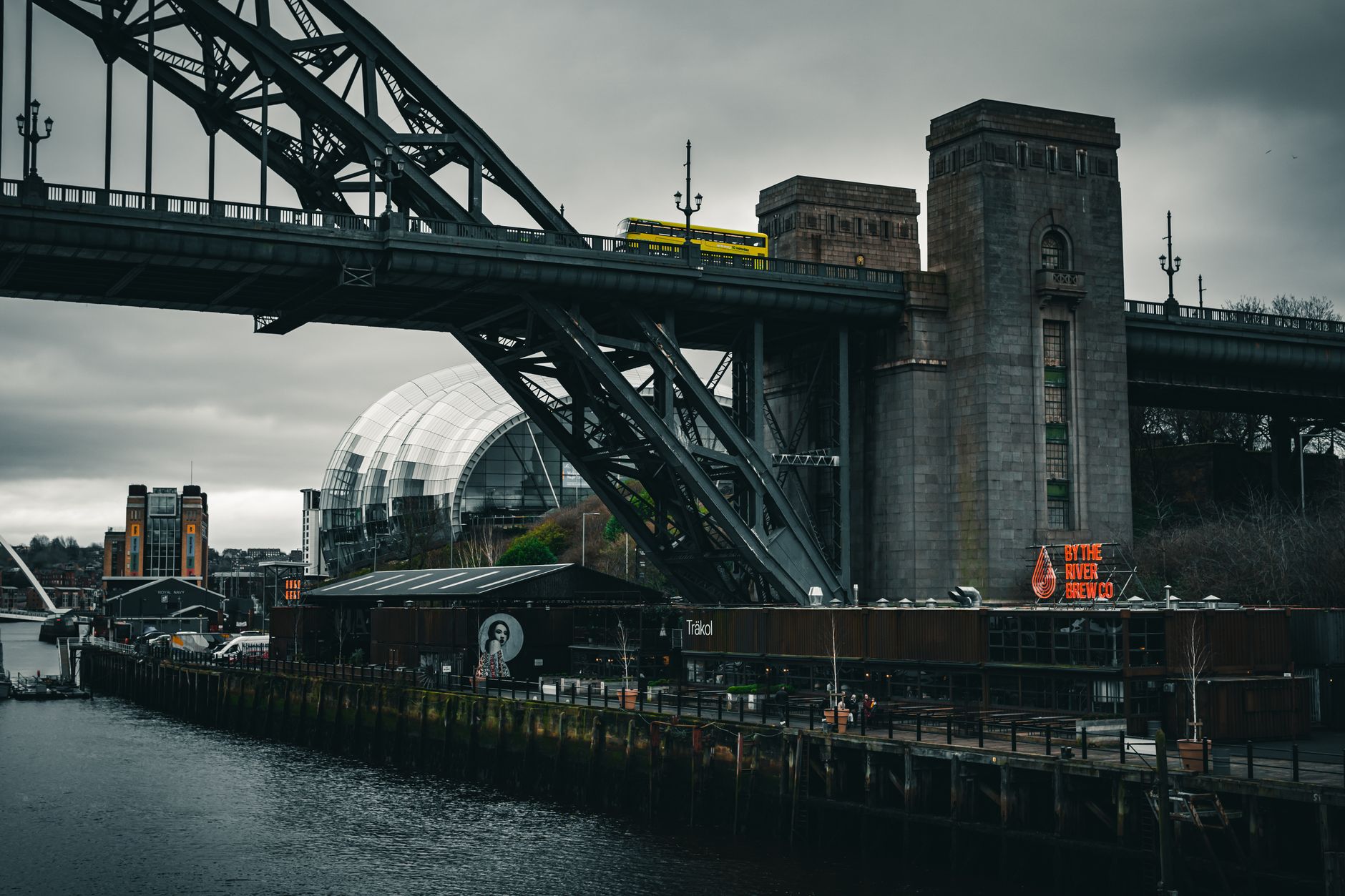Yellow bus crossing iconic urban bridge under overcast skies by the river.