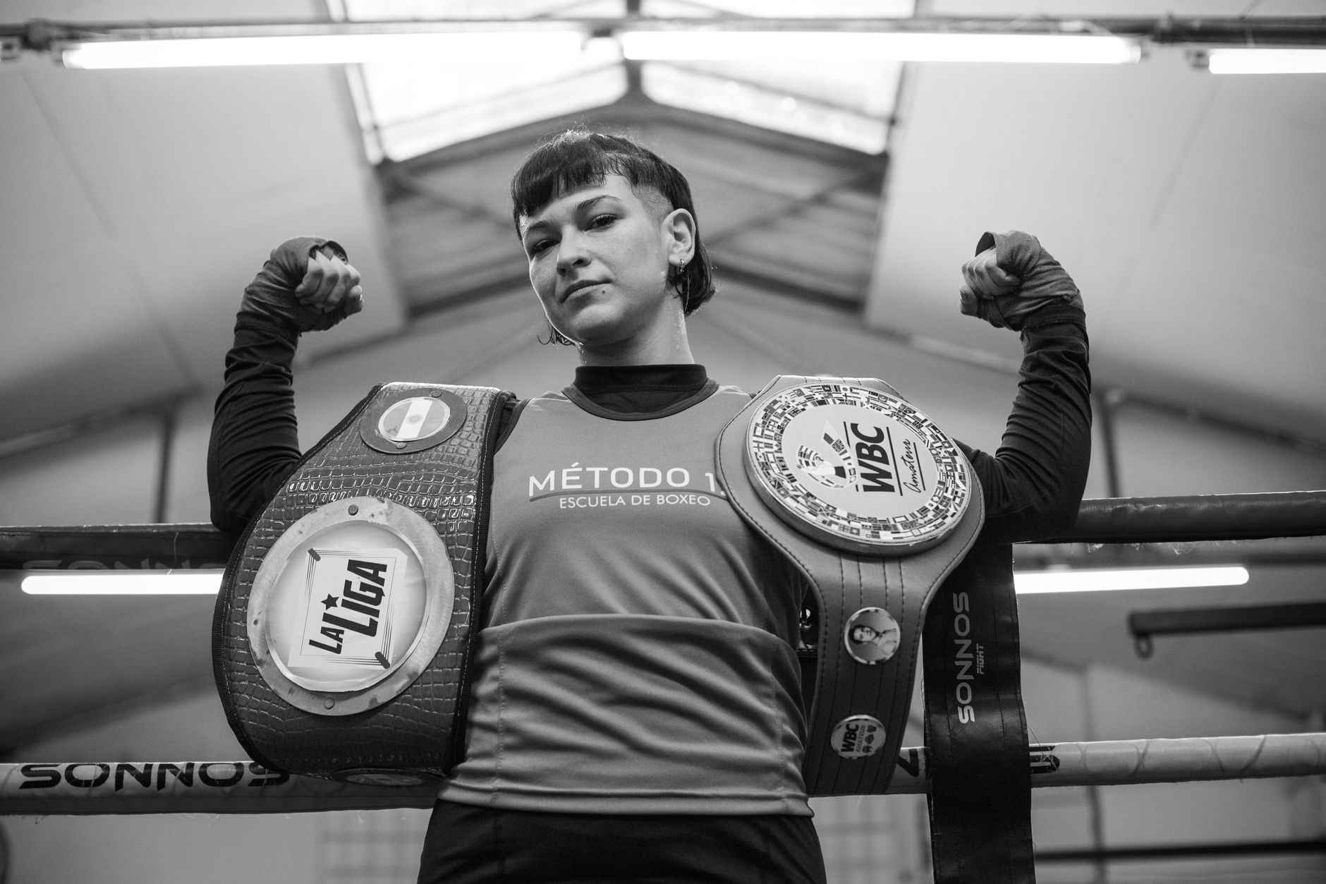 A female boxer proudly showcases her championship belts in a boxing gym.