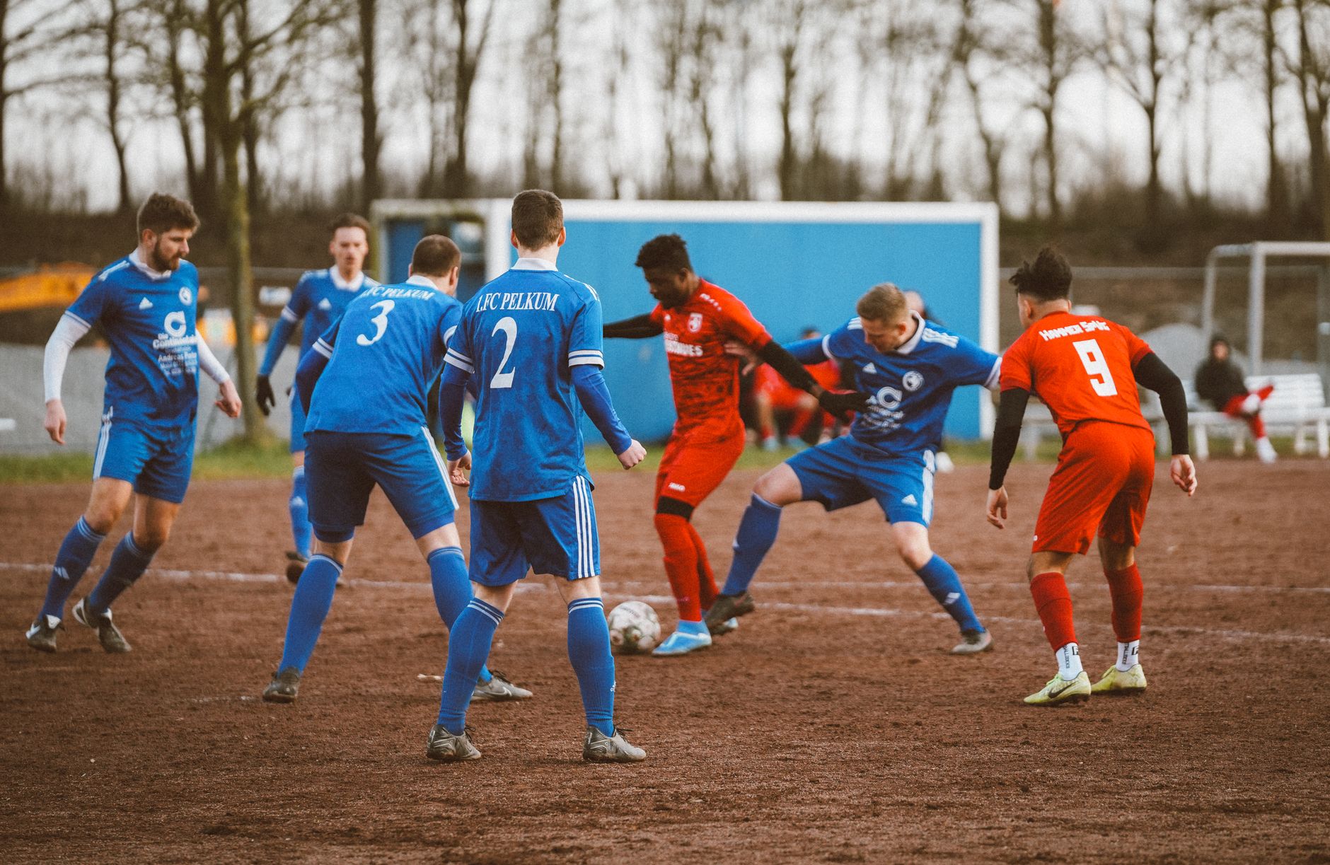 Intense amateur soccer match between blue and red team on a muddy field.
