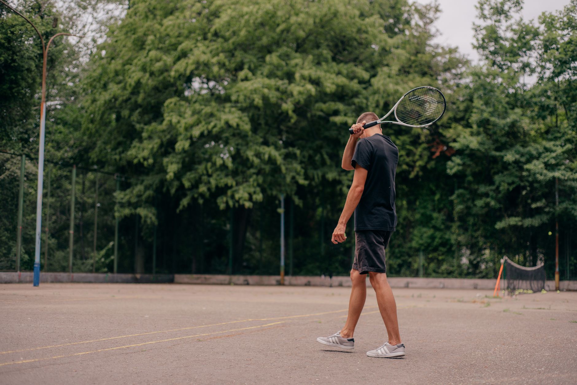 A young man holds a tennis racket while standing on an outdoor court surrounded by trees.