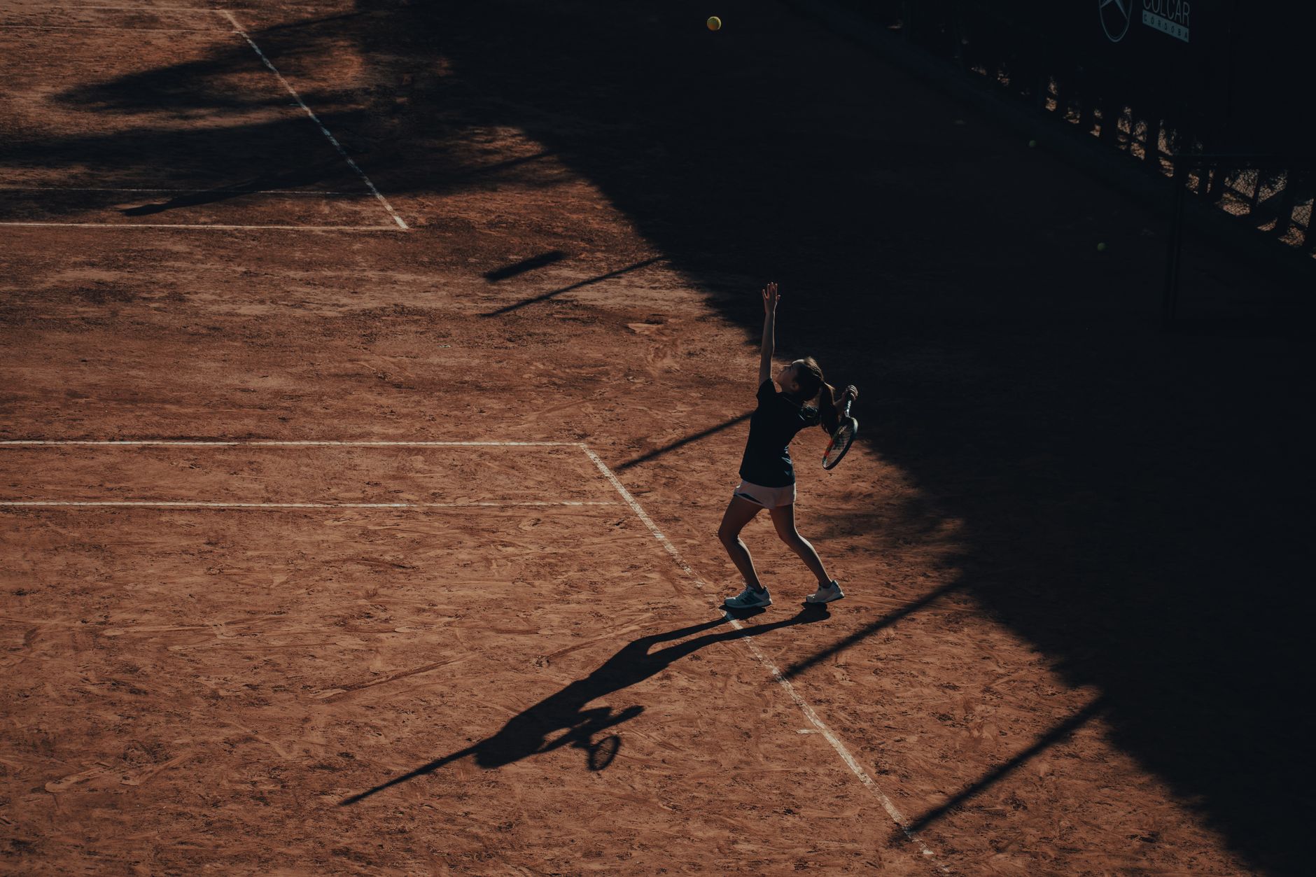 A tennis player serves on a clay court under dramatic lighting, highlighting shadows and motion.