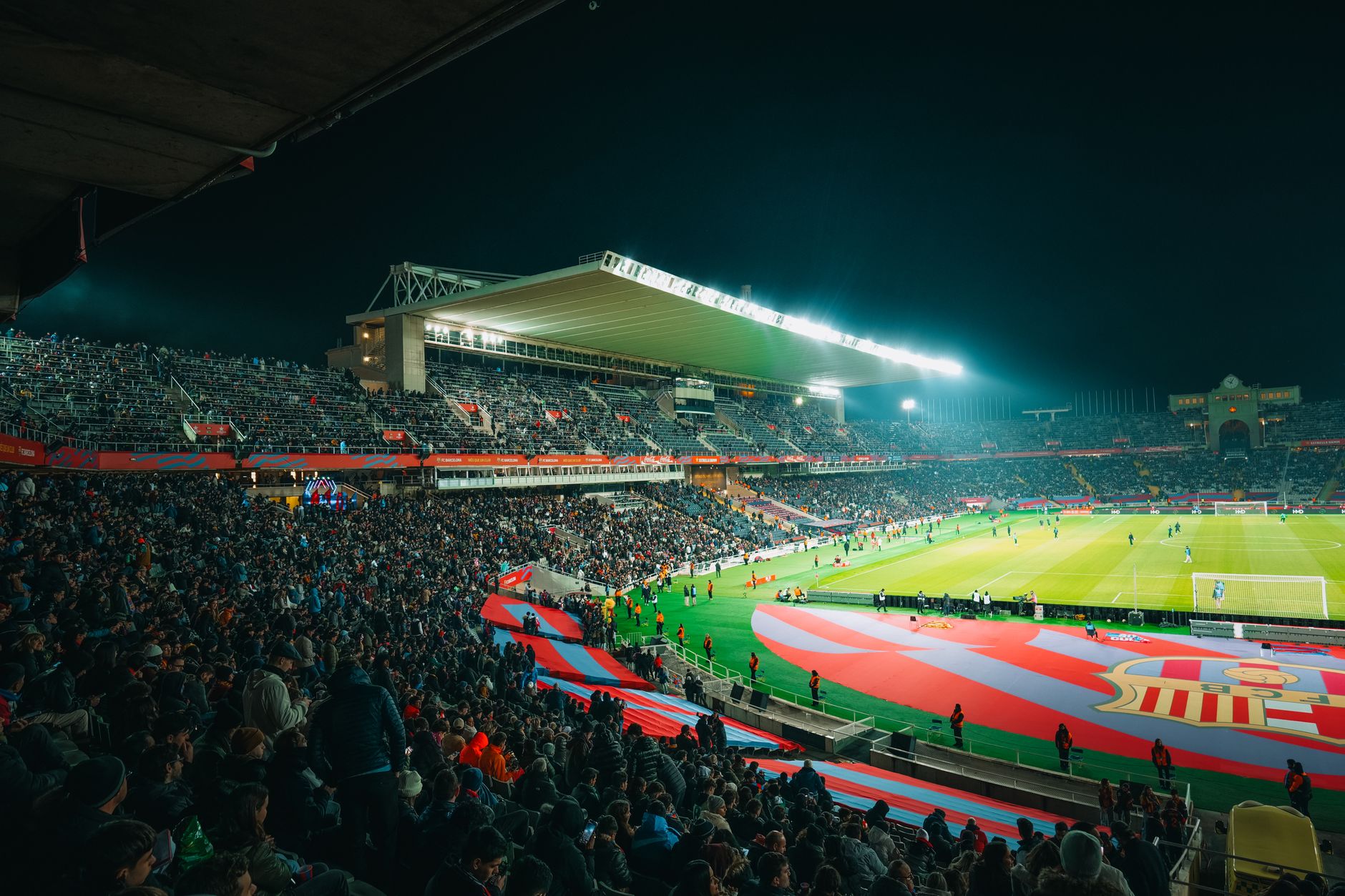 A crowded night-time football match in a brightly lit and bustling stadium, full of energy.