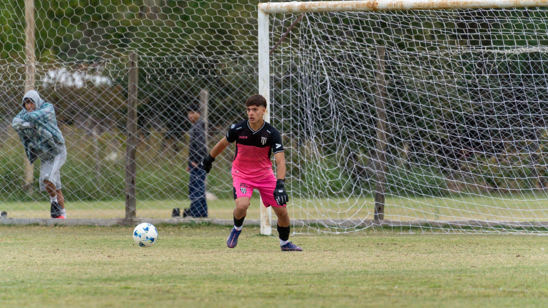 A young male goalkeeper in pink uniform guards the net during a soccer game outdoors on a grassy field.