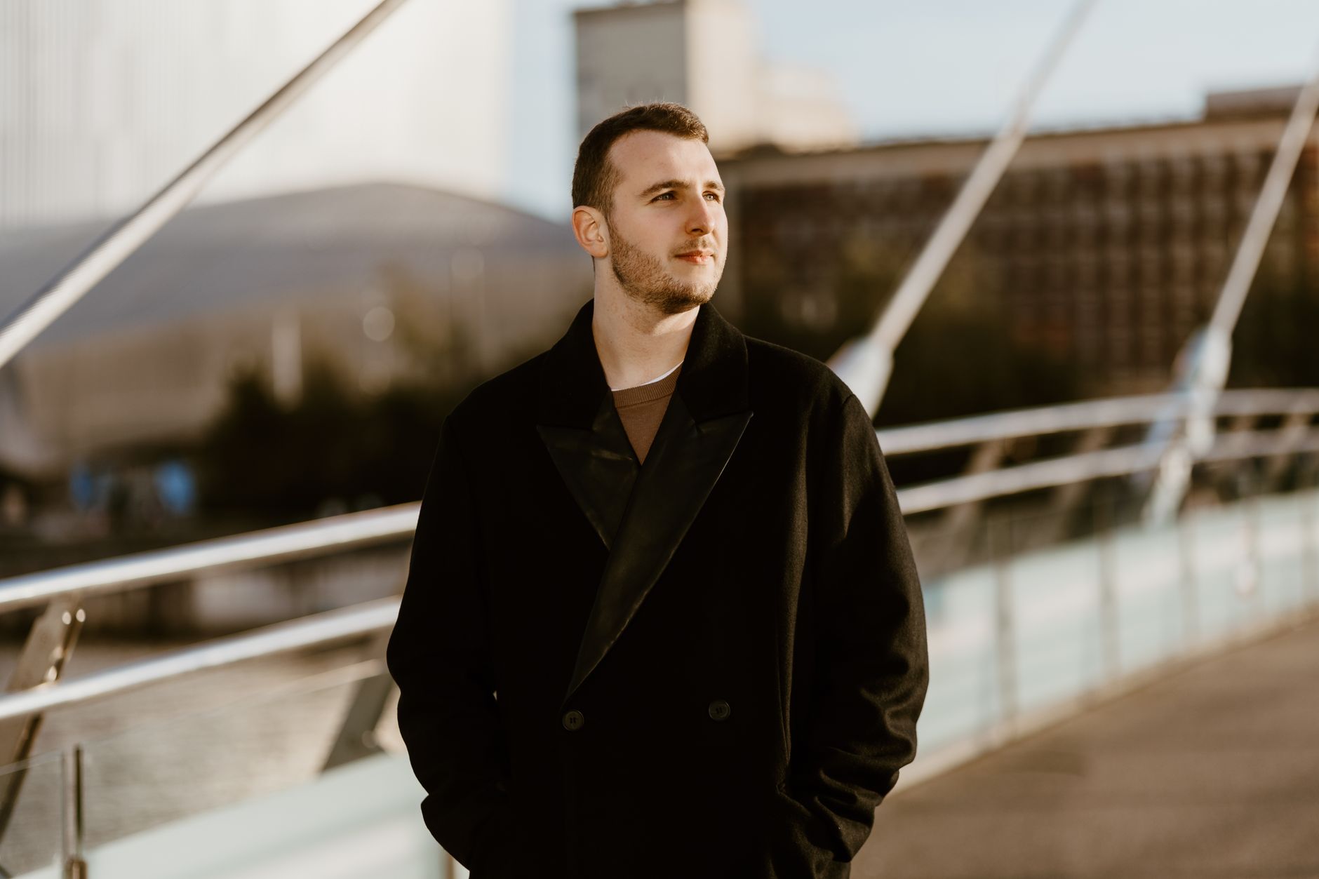Fashionable man in a coat standing on a modern bridge in Manchester during the day.