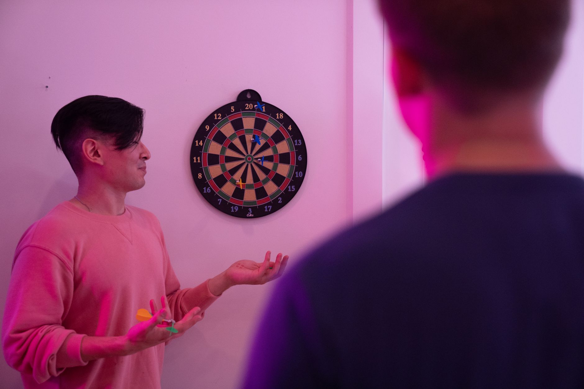 Two young men playing darts indoors with vibrant pink lighting, focusing on the dartboard.