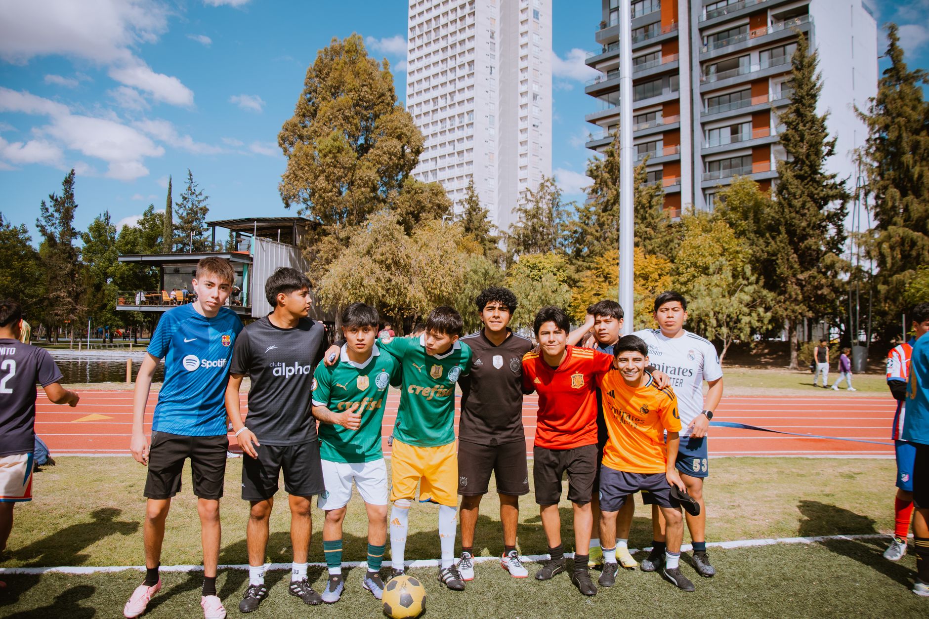Teenagers enjoy a soccer game outdoors on a sunny day, featuring diverse jerseys and urban background.