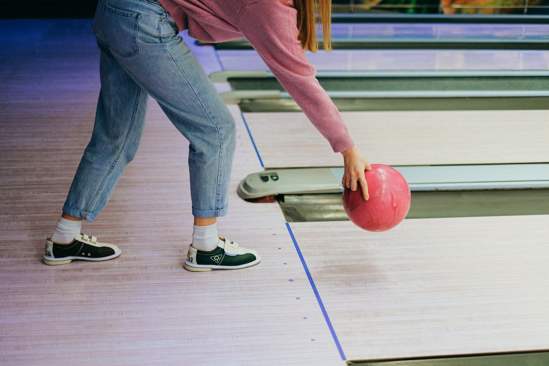 A woman in jeans and sweater playing bowling, throwing a pink ball down the lane indoors.