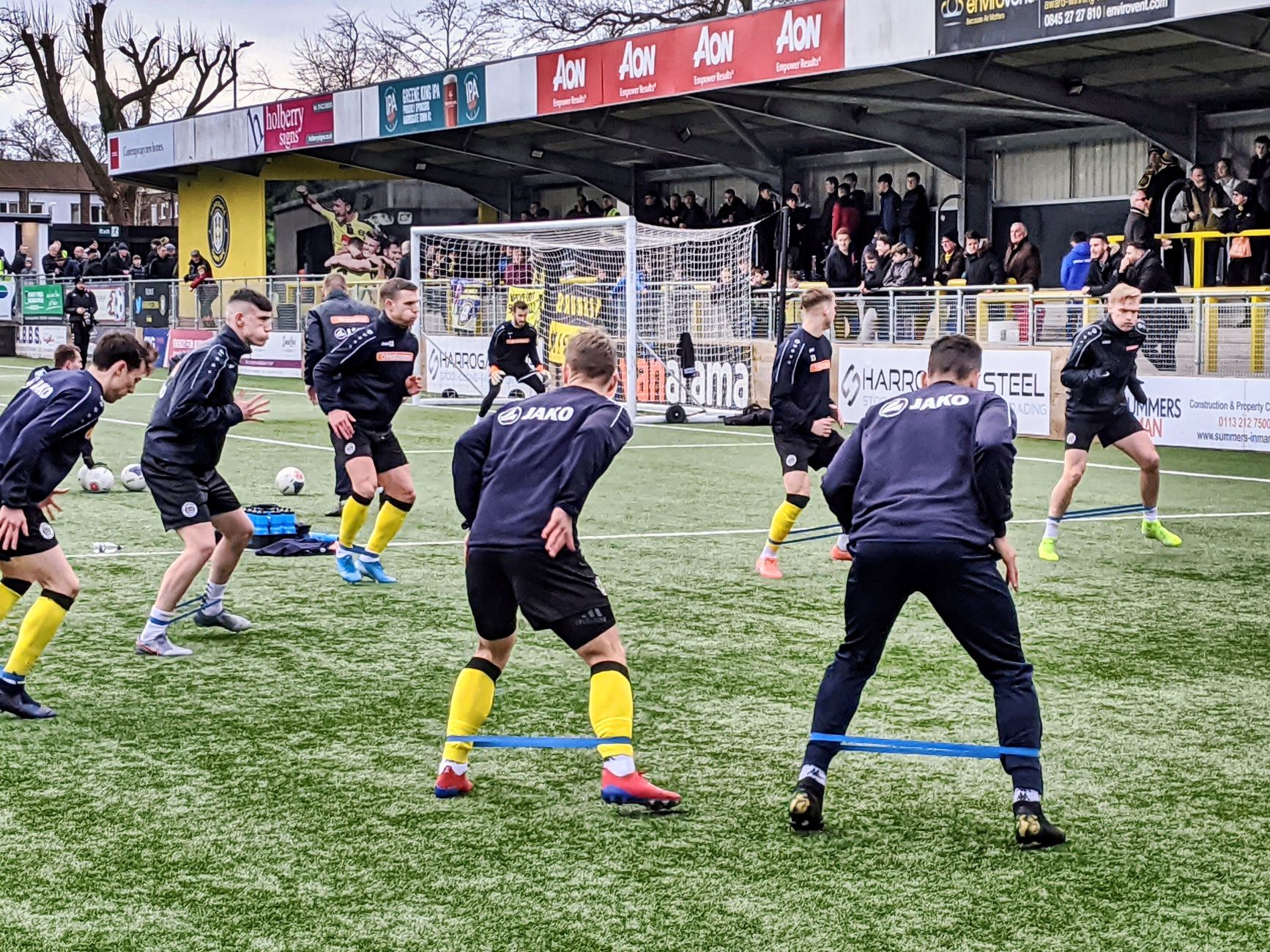 Players performing warmup exercises on a football field in England with spectators watching.
