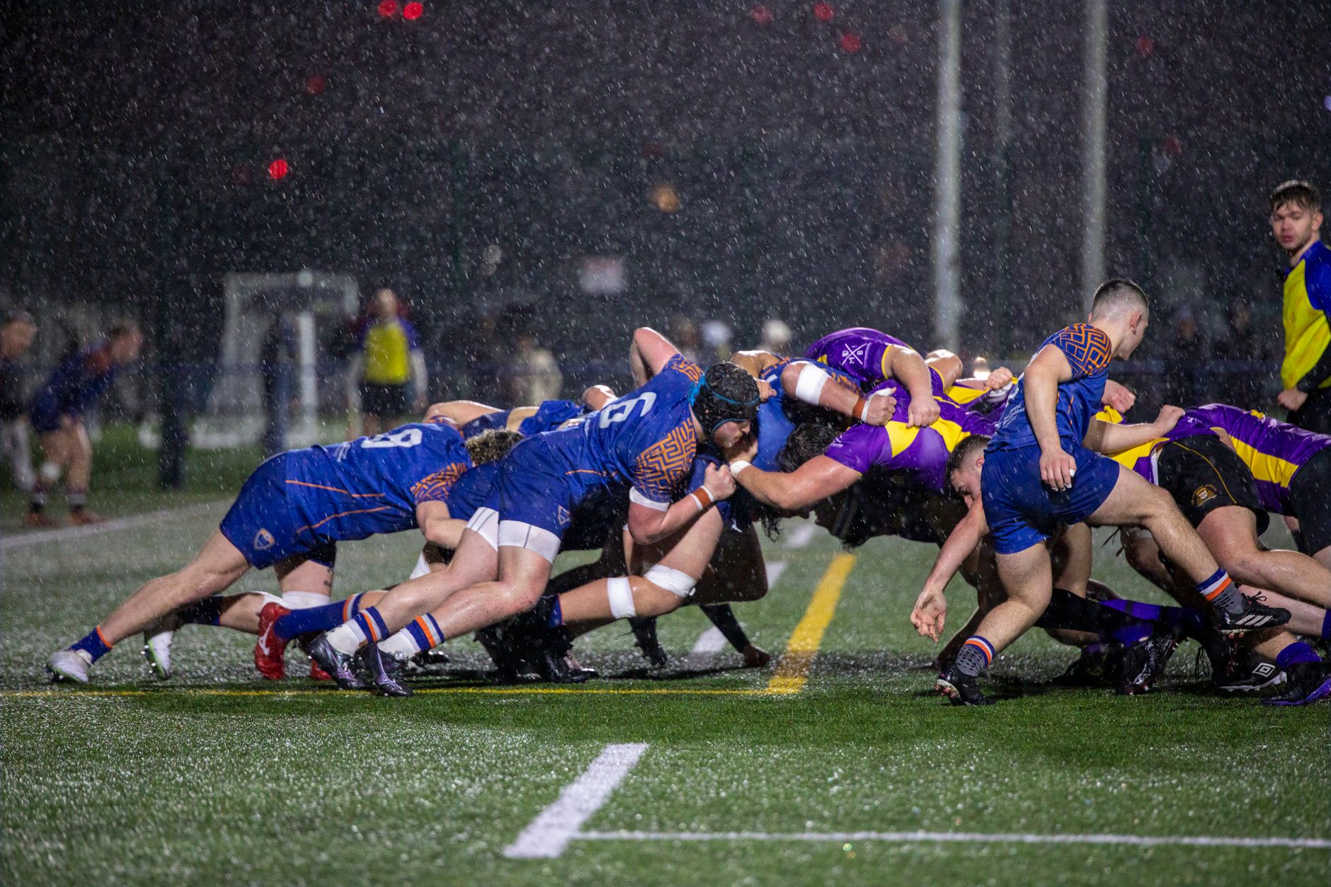 Dramatic rugby scrum at night in Manchester rain, showcasing teamwork and athleticism.