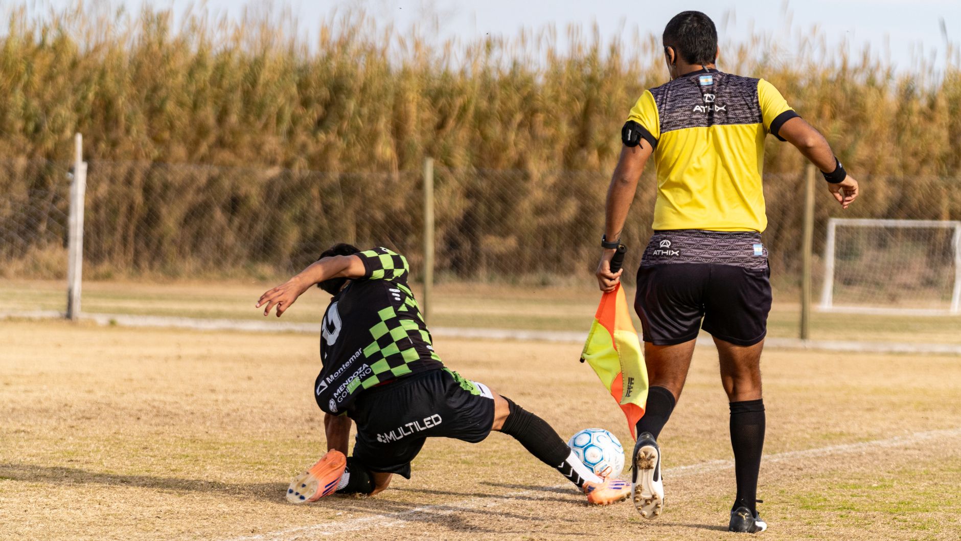 Soccer player tackles during a match as referee observes. Captured outdoors on a field.