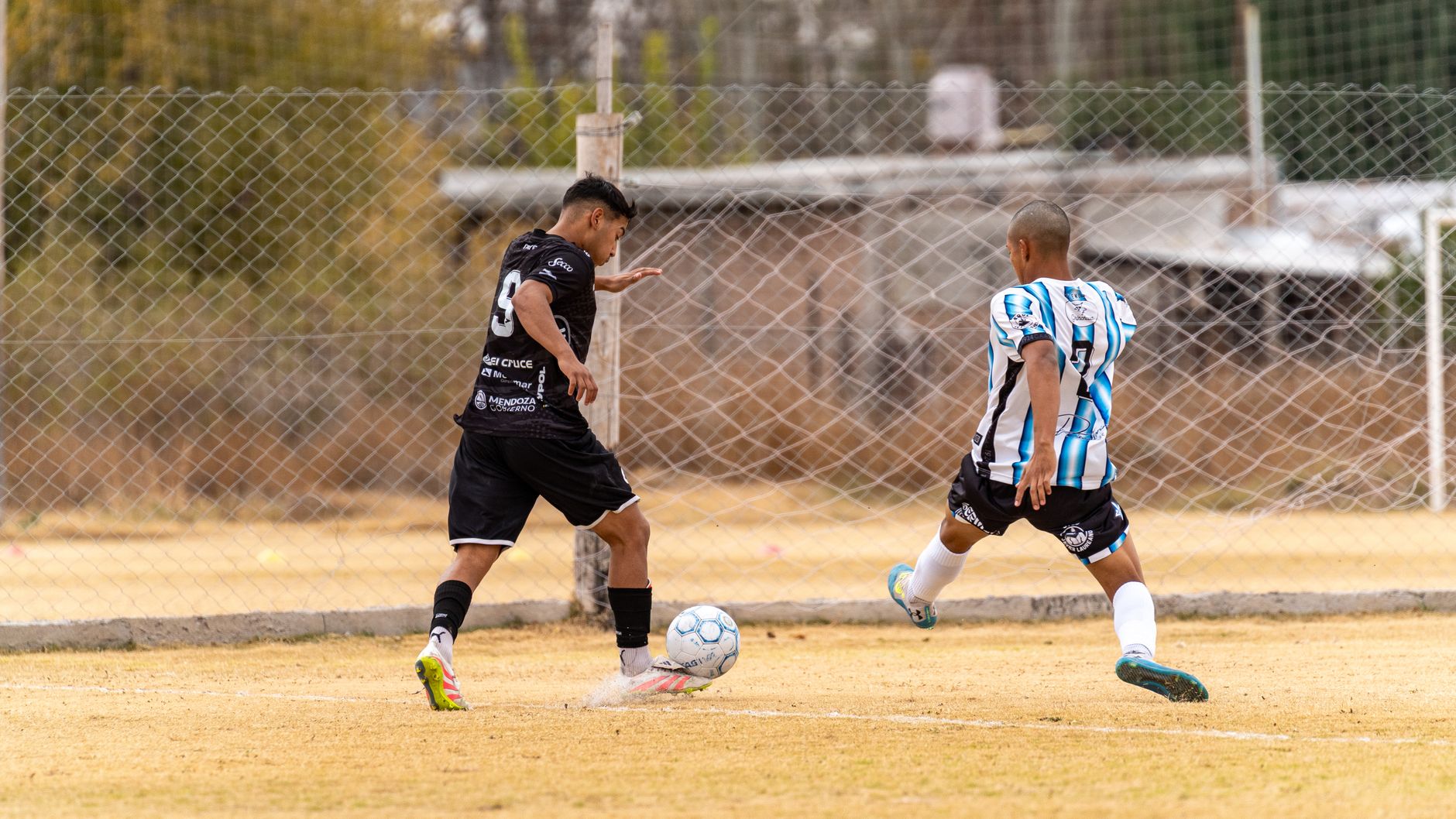 Two male soccer players in action on a grass field during a daytime match with chain-link fence backdrop.