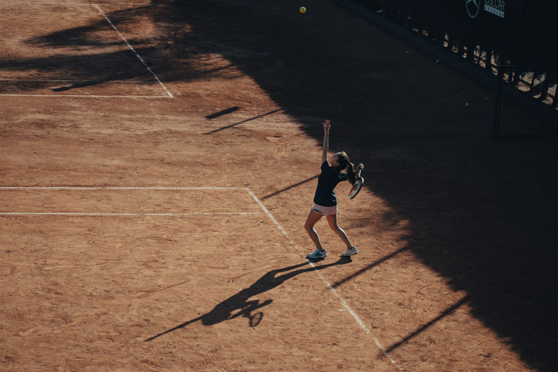 Aerial view of a woman playing tennis, serving on a clay court under sunlight.