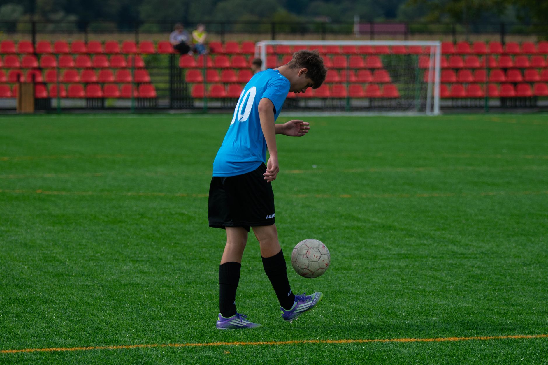 Teenage soccer player skillfully balancing a soccer ball on a grassy field with red stands.
