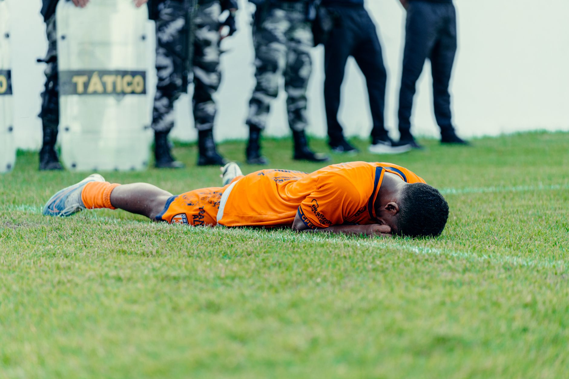 Injured football player in orange jersey lying on field with police presence.