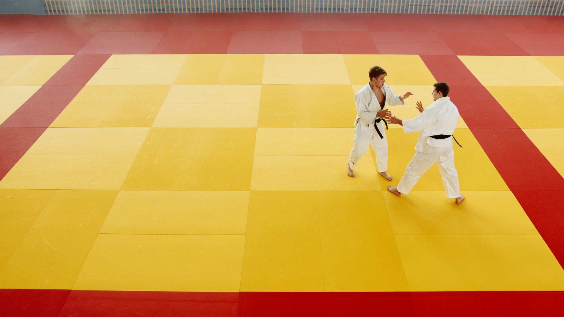 Two martial artists practice techniques on a yellow and red mat in an indoor gym.