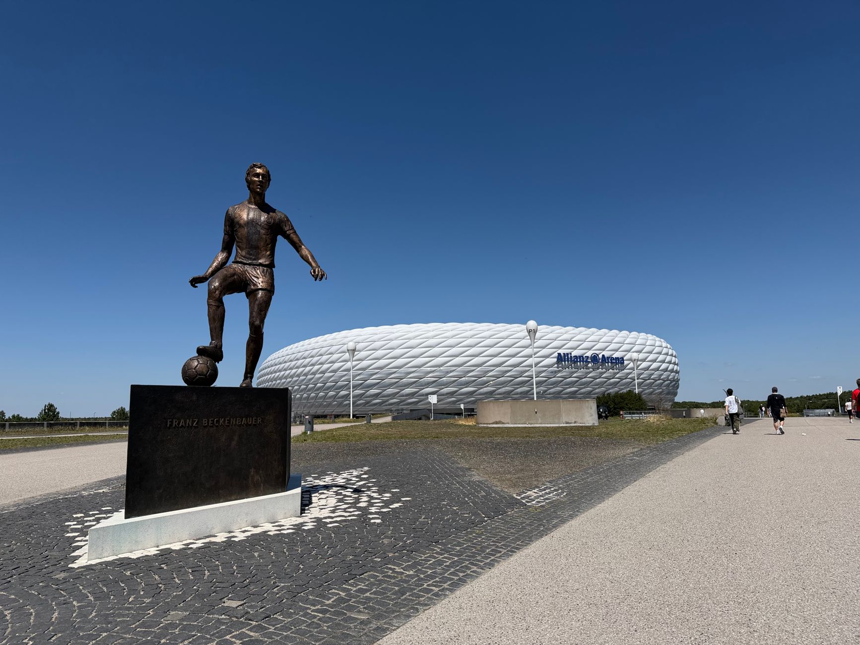 Statue of Franz Beckenbauer with Allianz Arena in Munich, Germany under a clear blue sky.